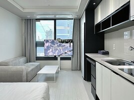 Habitación de hotel moderna con diseño minimalista, que incluye un sofá, una mesita, una zona de cocina con fregadero y armarios, y un ventanal con vistas a la ciudad.