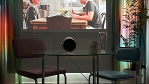 A living room with a television mounted on a wall, two chairs, a coffee table, and a potted plant.