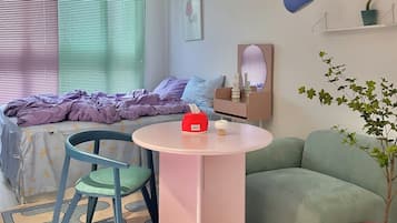 A cozy bedroom with a bed, a small round table, a chair, a green sofa, a potted plant, and a wall-mounted shelf.
