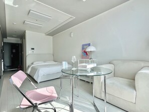 A modern hotel room with a bed, a glass table, a pink chair, a white sofa, and a painting on the wall.