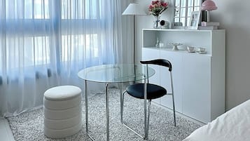 A modern bedroom with a glass table, a black chair, a white ottoman, and a bed.