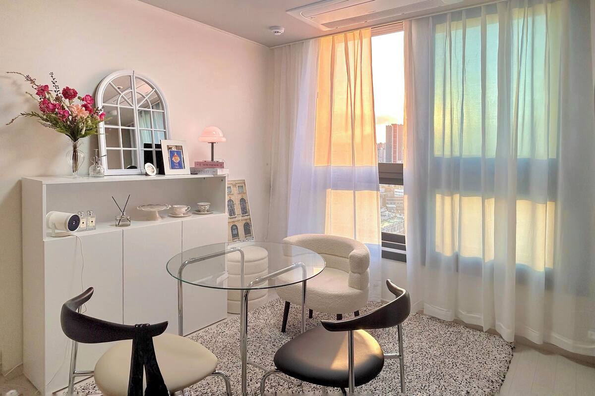 A modern hotel room with a glass table, two chairs, a white sofa, a white console, and a large window with sheer curtains.