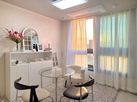 Una habitación de hotel moderna con una mesa de vidrio, dos sillas, un sofá blanco, una consola blanca y un gran ventanal con cortinas de gasa.