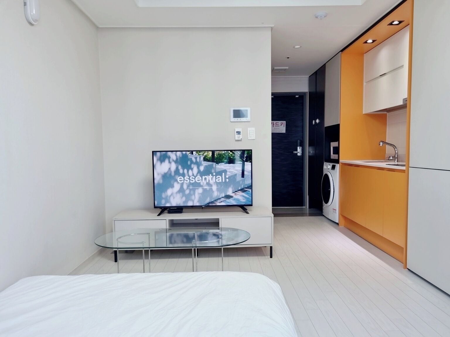Una camera d'albergo moderna con una TV a schermo piatto, un tavolino da caffè in vetro, un letto, un angolo cottura con lavatrice e una porta che conduce al bagno.