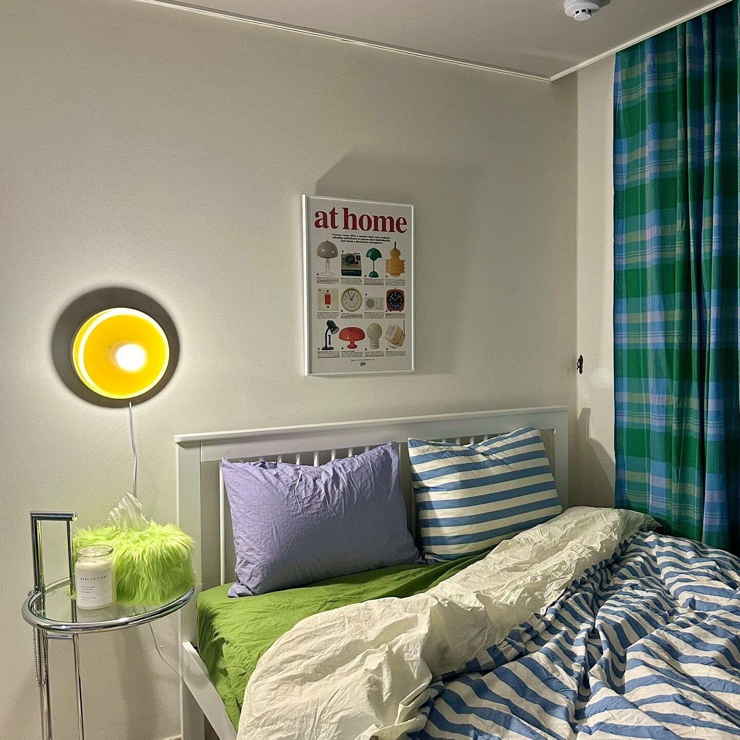 A bedroom with a bed, a bedside lamp, a poster on the wall, and a window with curtains.