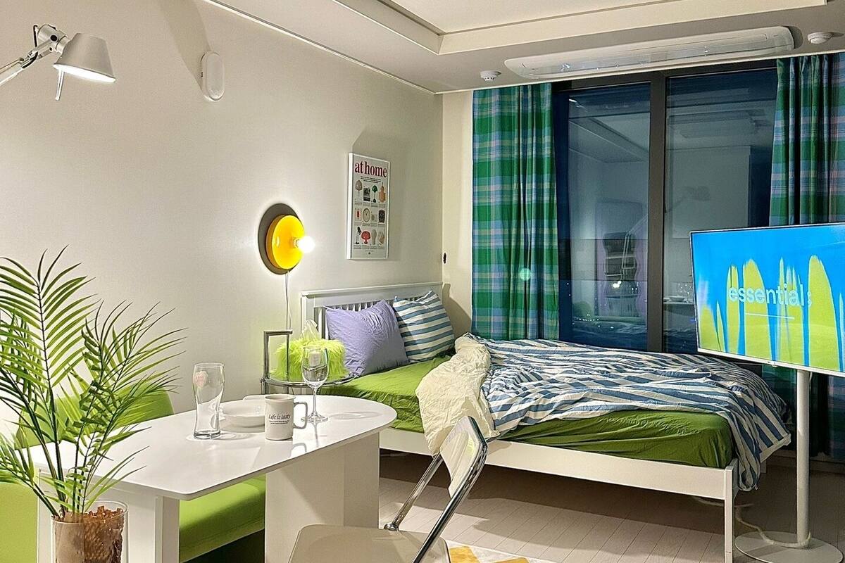 A modern bedroom with a bed, a desk, a chair, a potted plant, and a window with curtains.