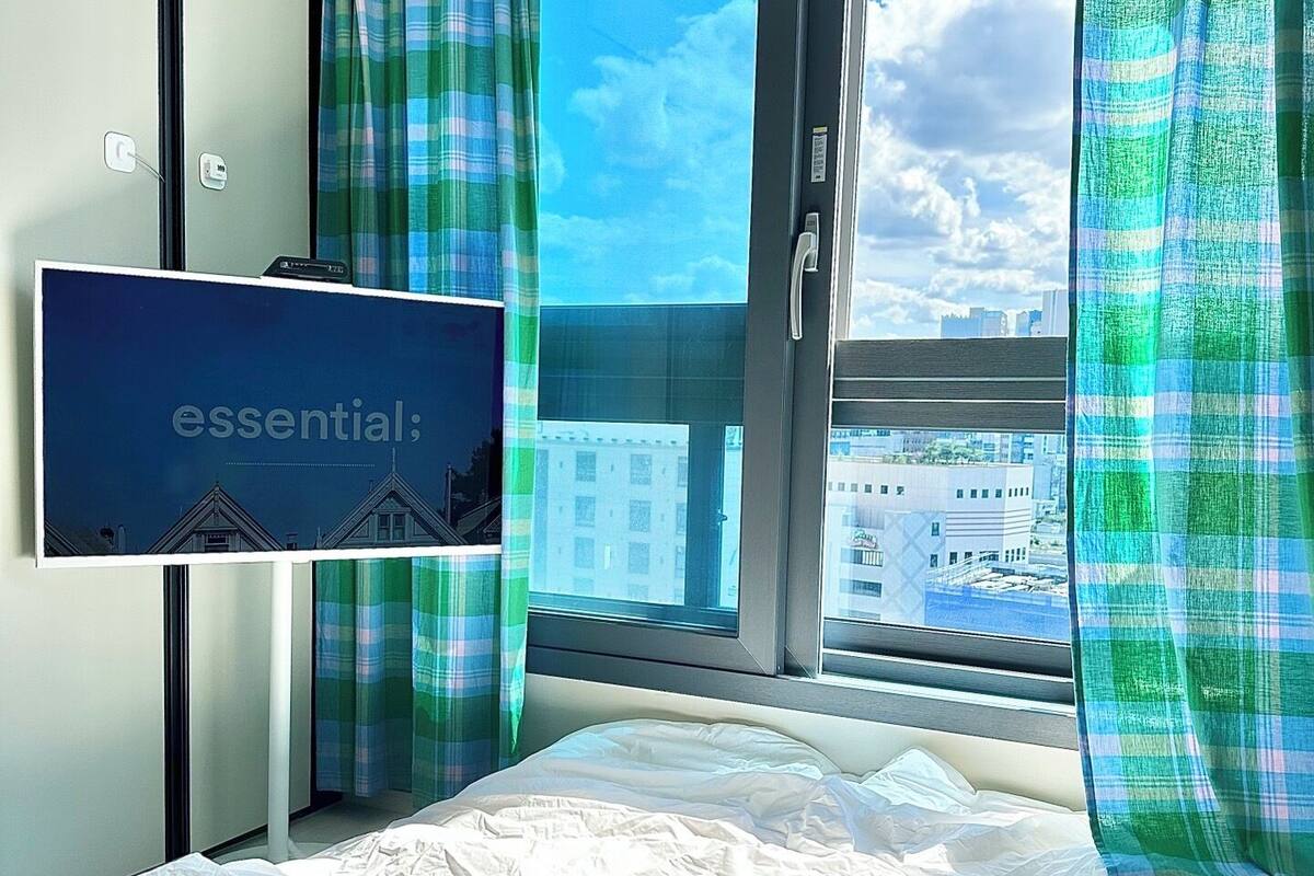 A hotel room with a bed, a TV on the wall, and a view of buildings through the window.