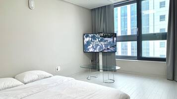 Una habitación de hotel moderna con una cama grande, un televisor de pantalla plana montado sobre una mesa de vidrio y vistas a la ciudad a través de amplios ventanales.