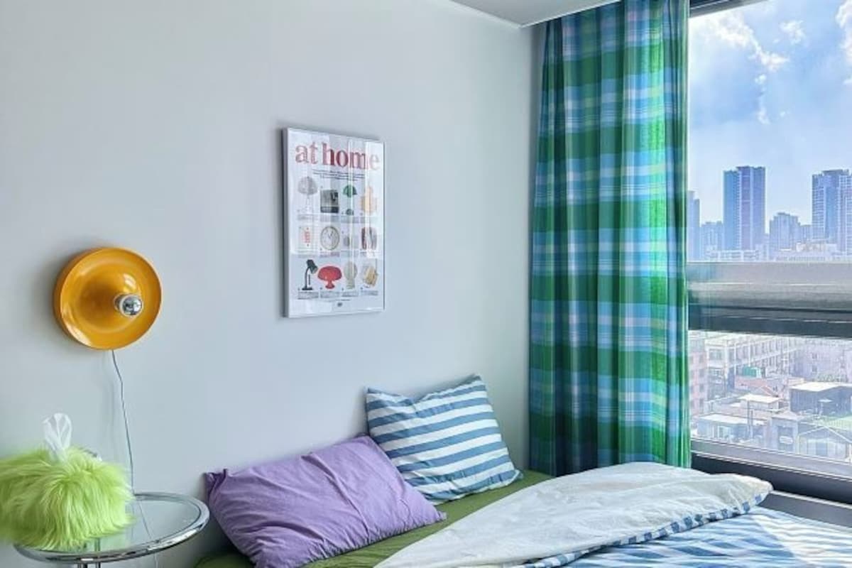 A bedroom with a bed, pillows, a poster, a lamp, and a view of a cityscape through a window with curtains.