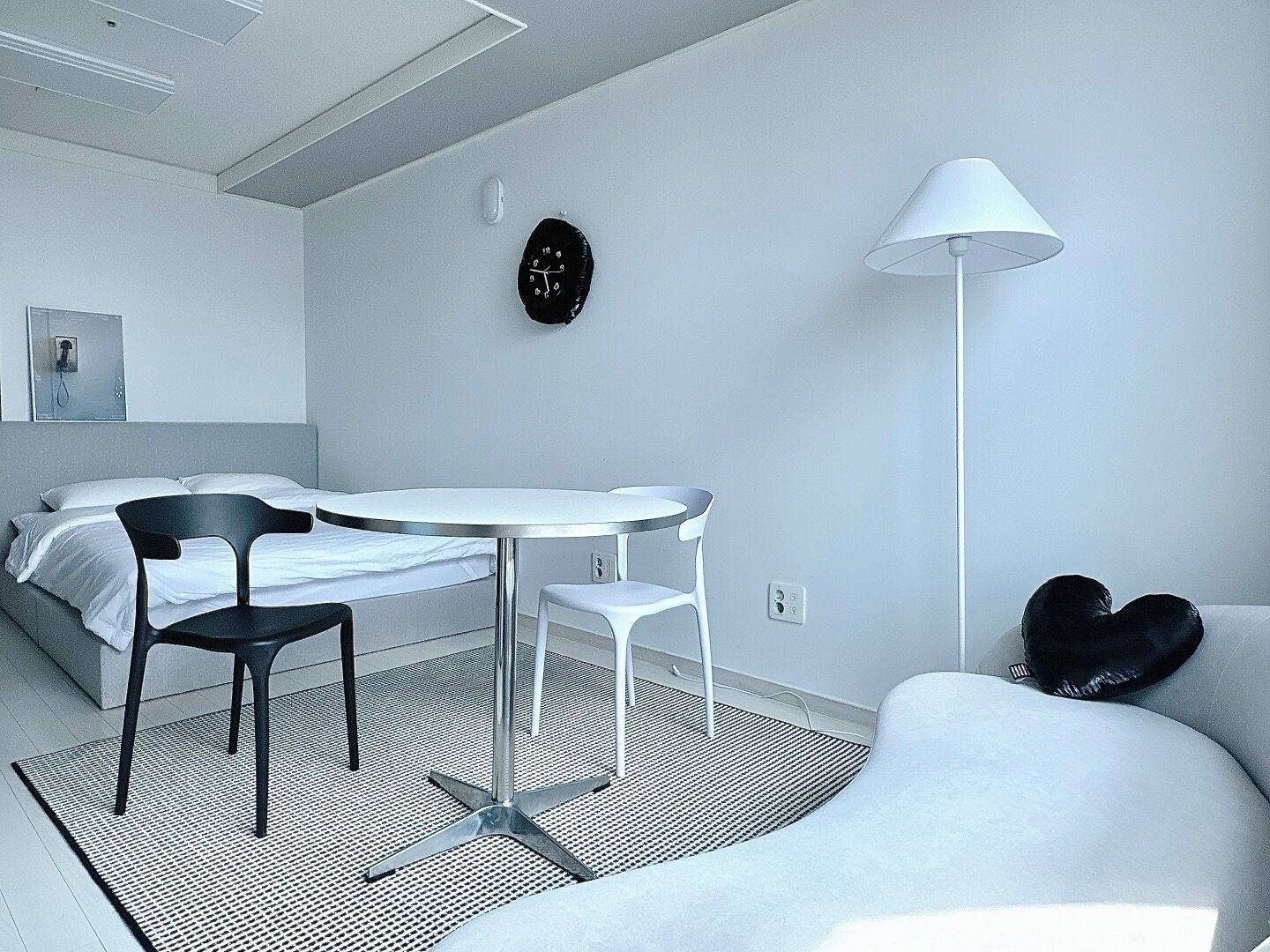 A modern, minimalistic room with a bed, a round table with two chairs, a white sofa, a floor lamp, and a wall clock.