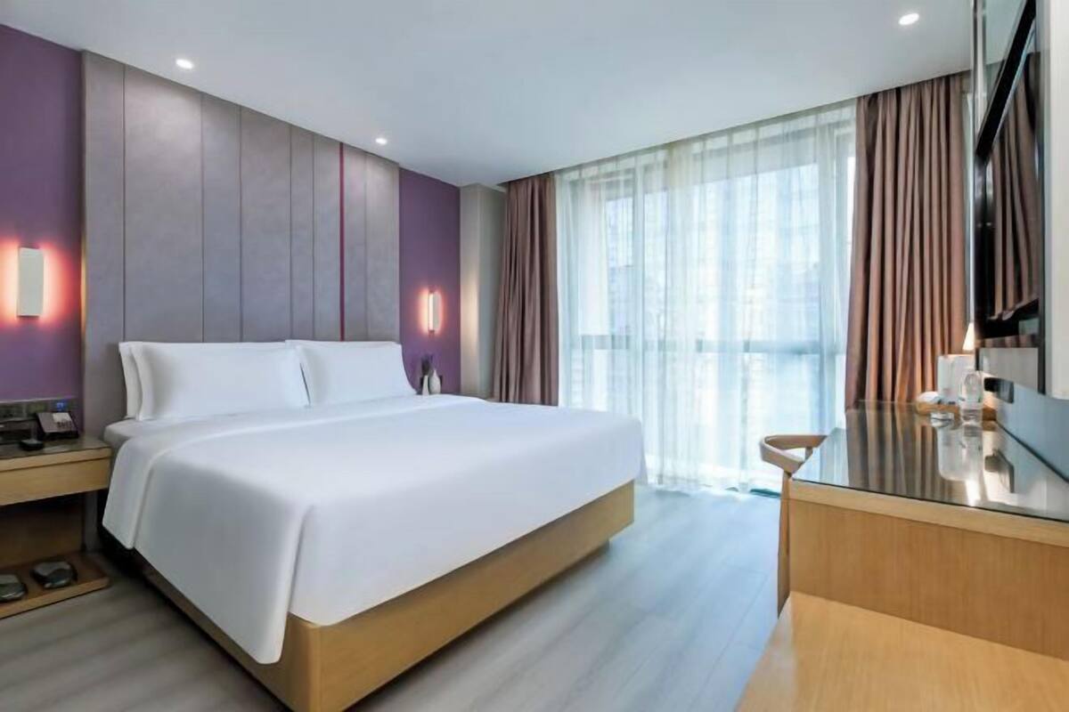 A modern hotel room with a large bed, a bedside table, a desk with a chair, and a large window with curtains.