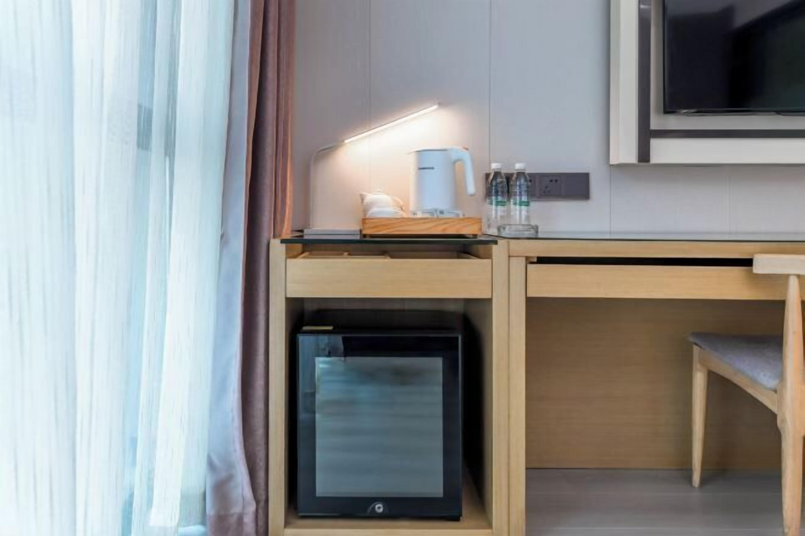 Kompakti hotellihuone, jossa on puinen työpöytä, sisäänrakennettu minijääkaappi, litteänäyttöinen televisio ja yöpöytä, jossa on lamppu.