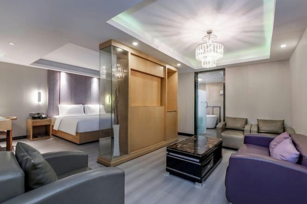 A modern hotel room with a bed, a seating area with sofas, a coffee table, and a large mirror.