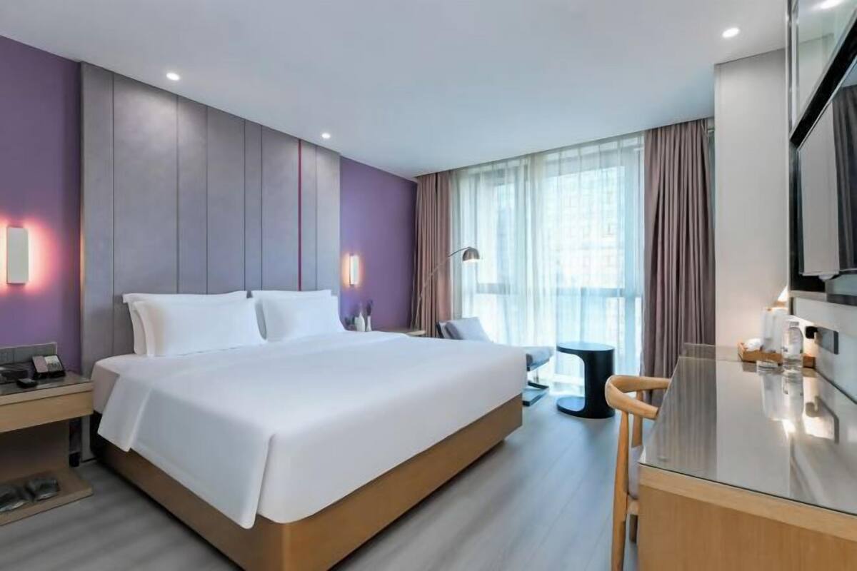 A modern hotel room with a large bed, bedside table, lamp, and a view of the city through the window.