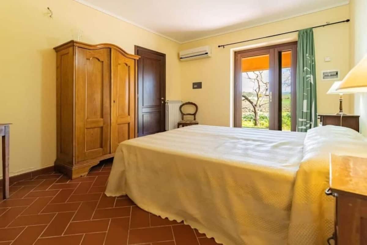 Quarto com cama, armário de madeira, radiador, janela com vista e porta.
