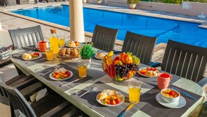 A poolside dining area with a table set for breakfast, including fruits, bread, and beverages. - Villa Paloma (Albufeira)