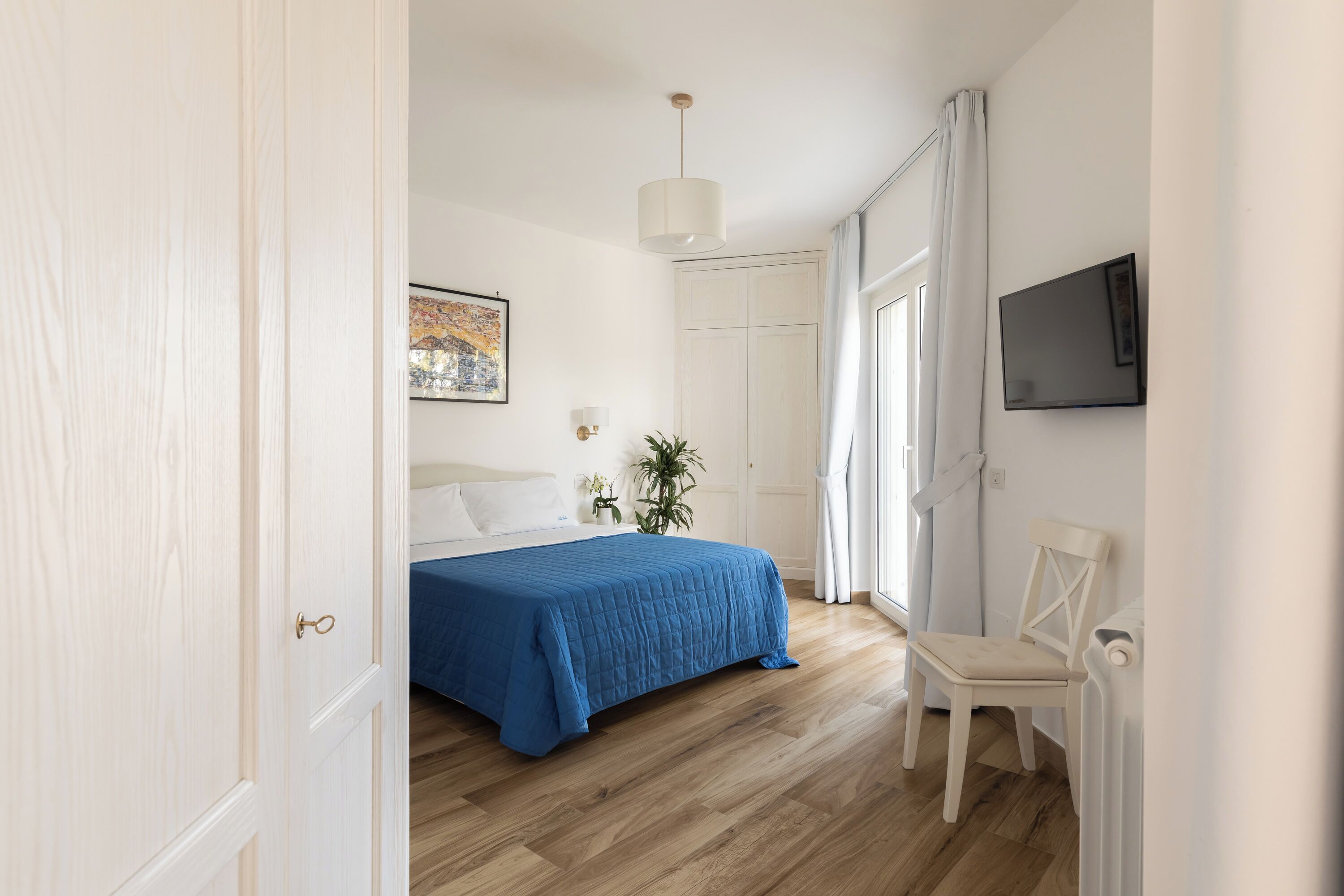 Un dormitorio con una cama, una silla, un televisor y un ventanal con cortinas.