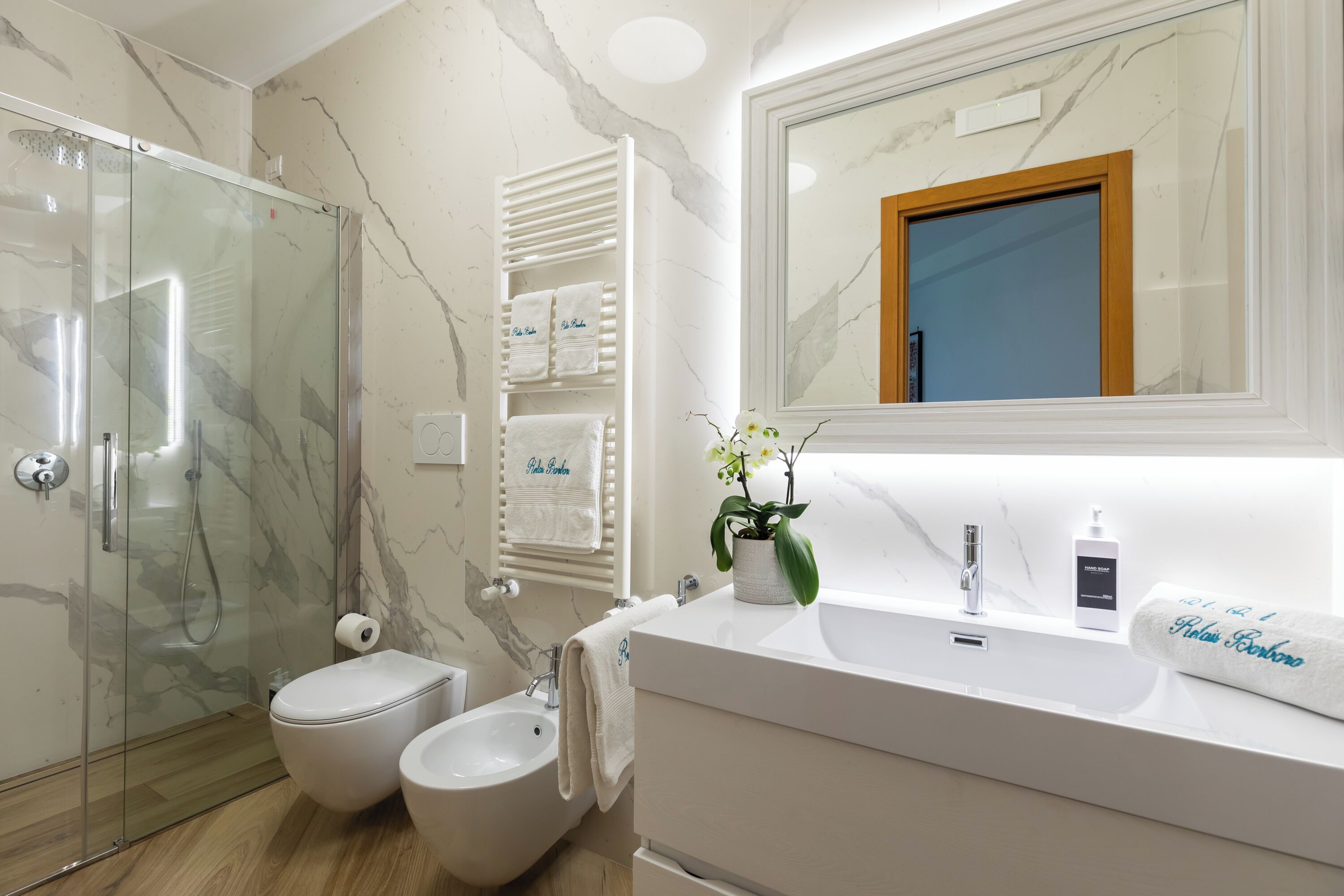A modern bathroom with a glass shower enclosure, a bidet, a sink with a mirror, and a towel rack with multiple towels.