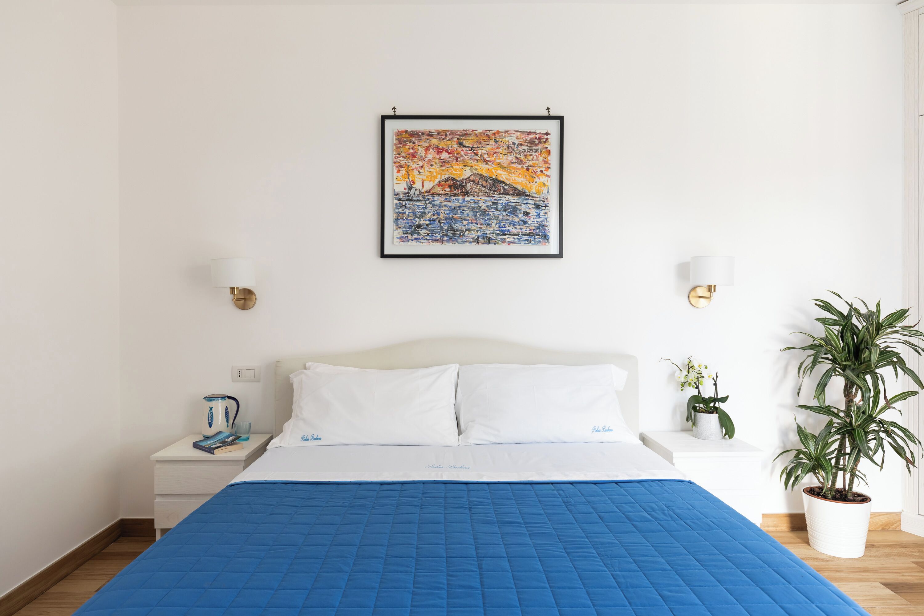 Un lit bien fait, avec une couette bleue, une tête de lit blanche et une œuvre d’art encadrée au-dessus.