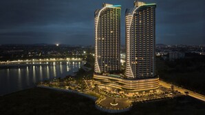 Dos edificios altos de diseño curvo, rodeados por un área ajardinada con una carretera y una masa de agua.