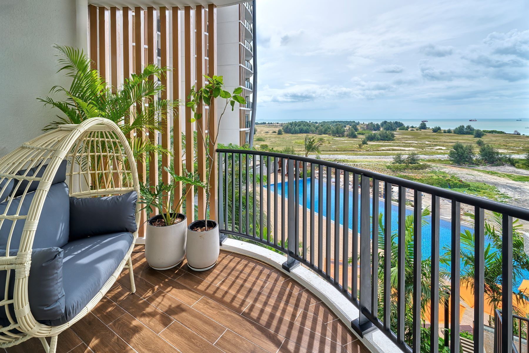 Un balcón con una silla de mimbre, dos plantas en macetas, vista a una piscina y paisaje distante.