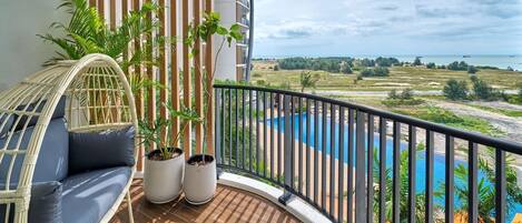 Un balcón con una silla de mimbre, dos plantas en macetas, vista a una piscina y paisaje distante.