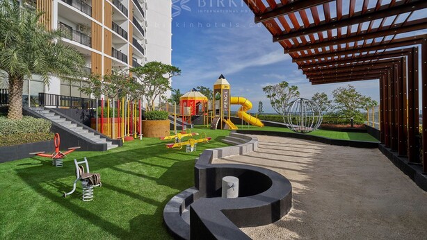 Zona de juegos infantiles con toboganes y columpios, un elemento circular de hormigón, y un edificio con balcones y una pérgola de madera.