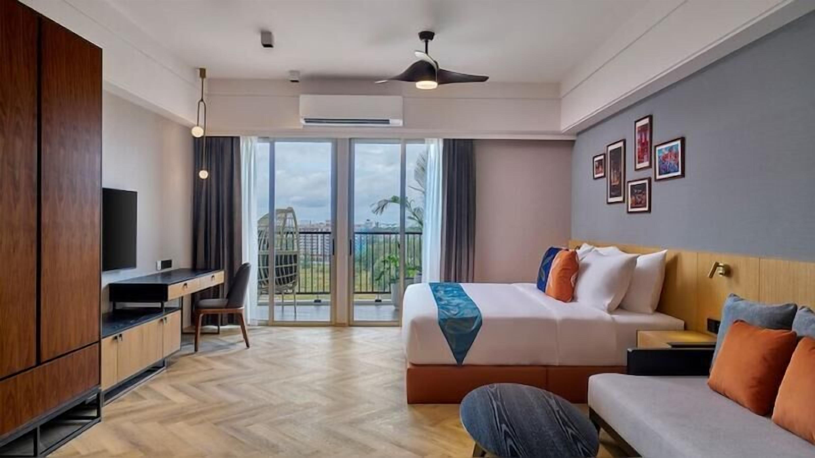 A hotel room with a bed, a desk, a sofa, a balcony with a view, and a ceiling fan.