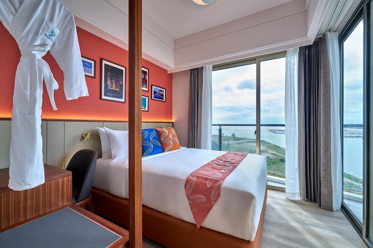 A hotel room with a bed, a desk, a chair, a robe, and a large window with a view of the sea.