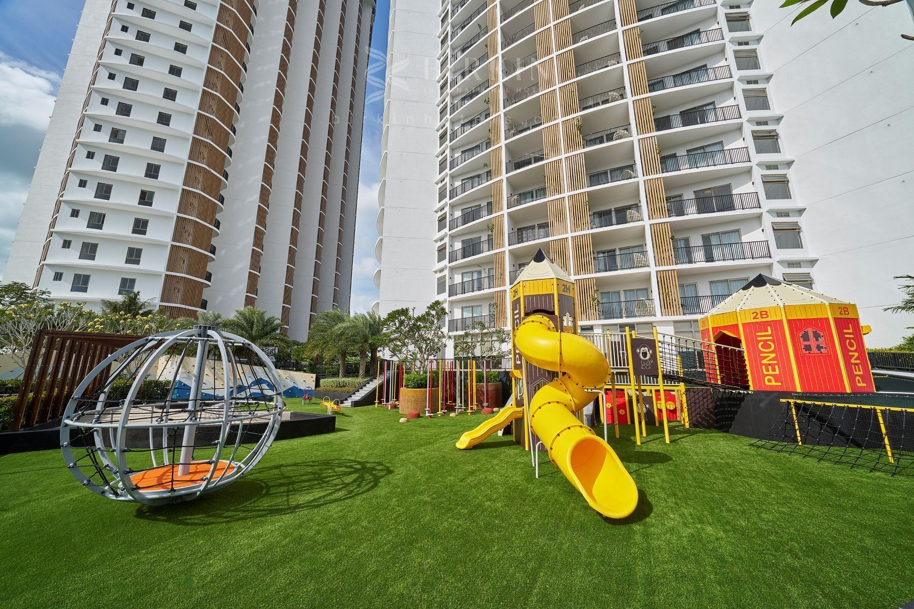 A playground with a slide and swings, surrounded by artificial grass, in front of a high-rise building.