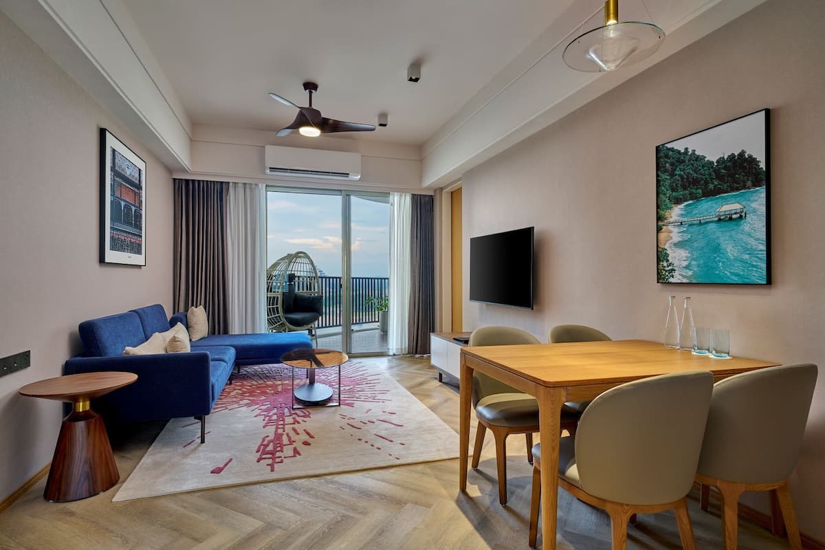 A modern hotel room with a blue sofa, a wooden dining table with chairs, a large TV, and a balcony with a view of the sea.