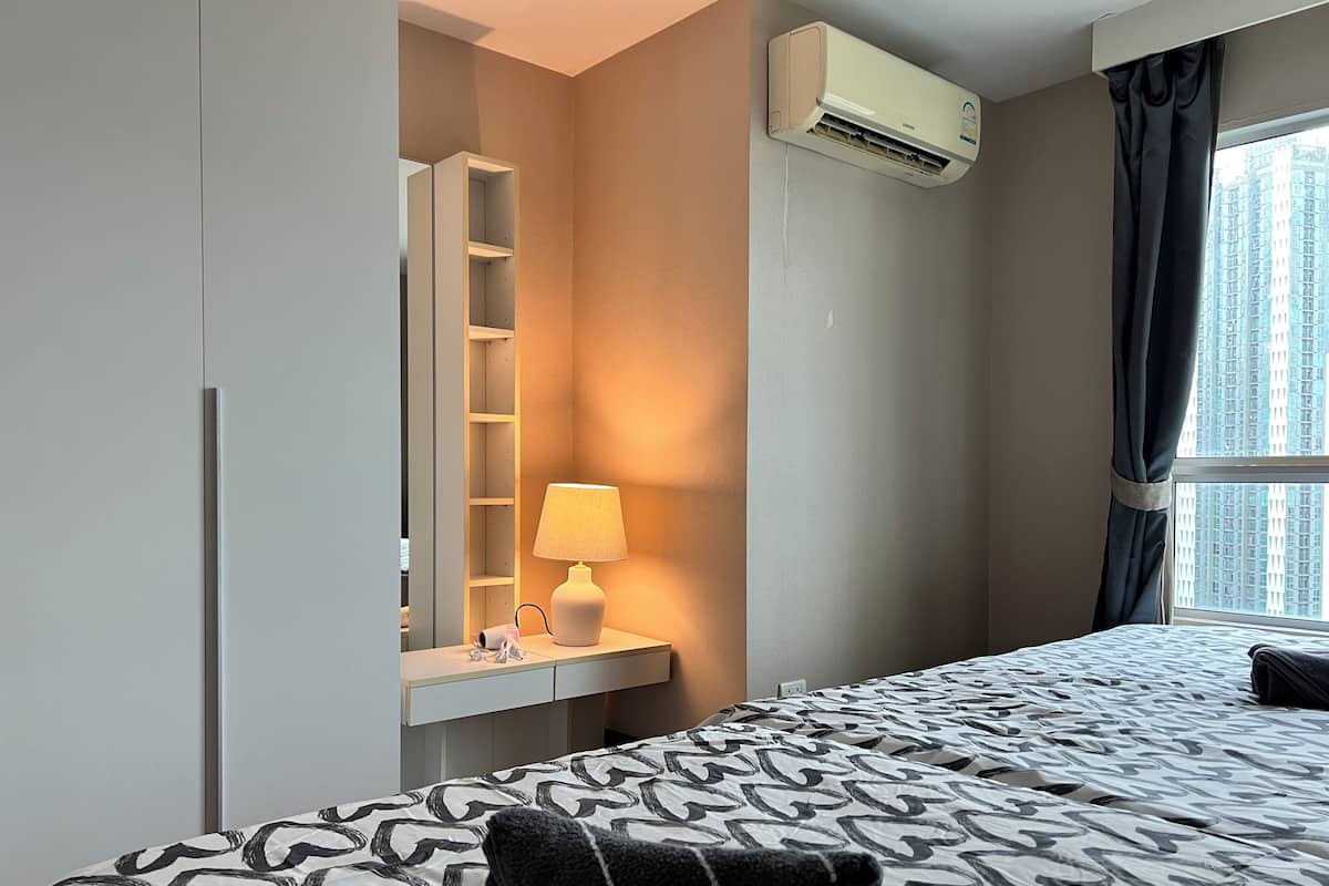 A hotel room with a bed, a bedside lamp, an air conditioning unit, a window with curtains, and a wardrobe.