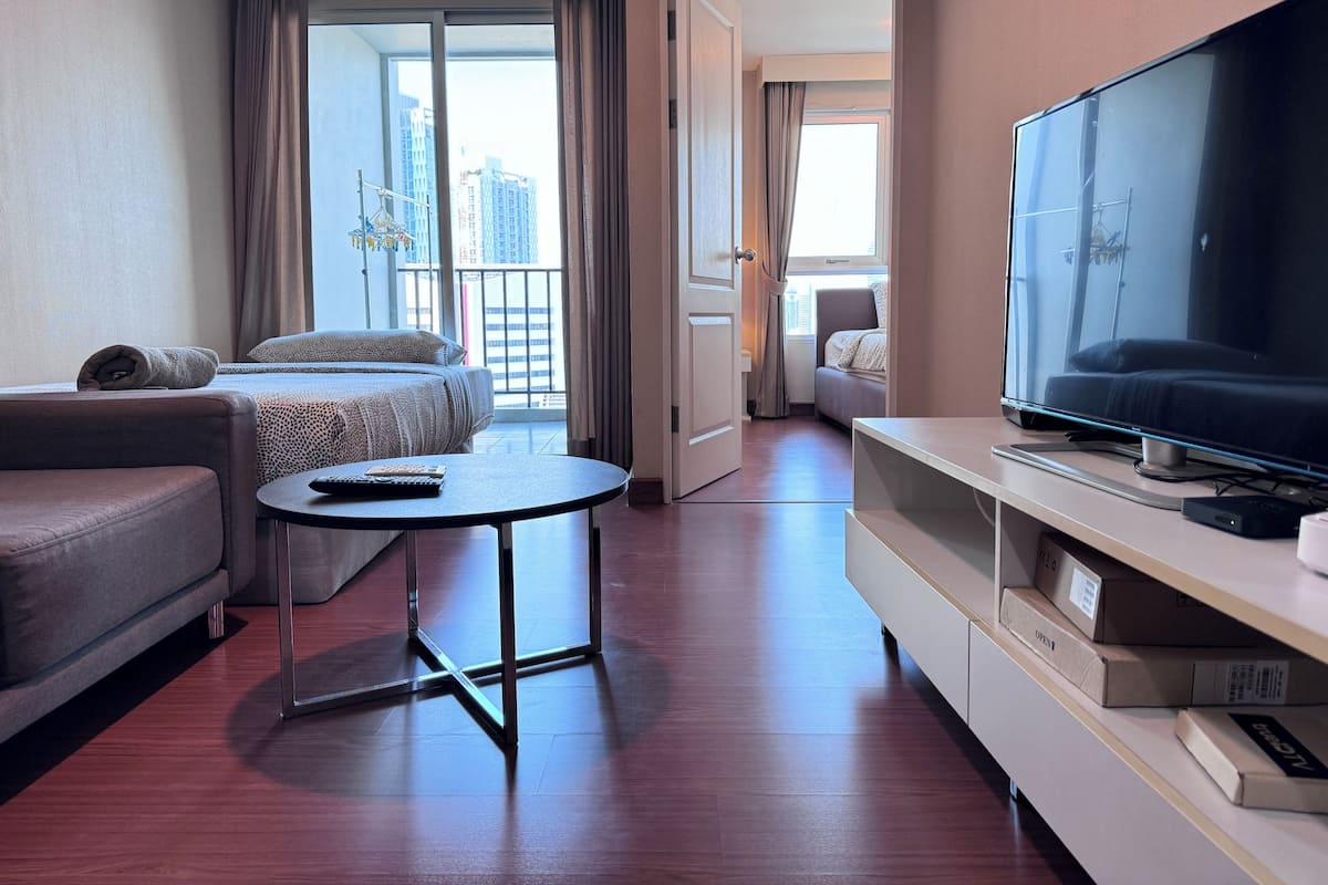 A hotel room with a bed, a sofa, a TV, a small table, and a balcony with a city view.