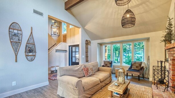 Ein geräumiges Wohnzimmer mit einem beigefarbenen Sofa, einem Holztisch und einem Kamin. Eine Treppe führt auf eine obere Ebene, und große Fenster sorgen für natürliches Licht.