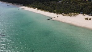 Eine Luftaufnahme eines Strandes mit klarem türkisblauem Wasser, einem sandigen Ufer und einem dicht bewaldeten Hügel im Hintergrund mit verstreuten Gebäuden.