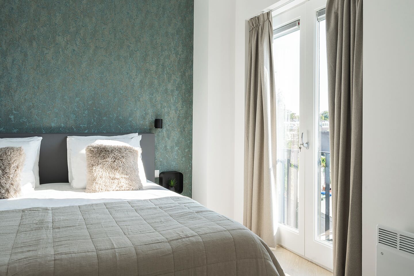 Ein Schlafzimmer mit Bett, Kopfstütze, Nachttisch, Fenster mit Vorhängen und Blick nach draußen.