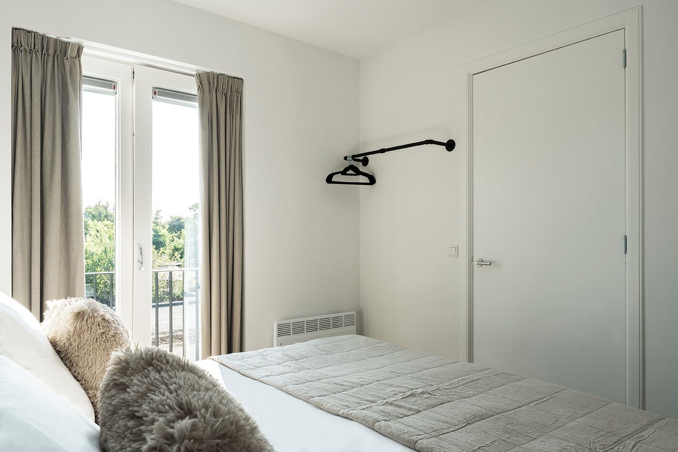 Un dormitorio con cama, ventana con cortinas, radiador y una puerta.