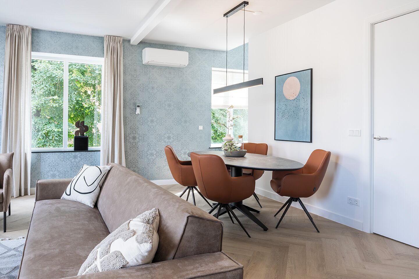Ein modernes Wohnzimmer mit einem grauen Sofa, einem Esstisch mit orangefarbenen Stühlen und einem großen Fenster mit Vorhängen.