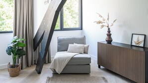 A modern living room with a grey sofa, a wooden floor, a large window with curtains, a potted plant, a woven rug, a wooden console table, and a framed picture.