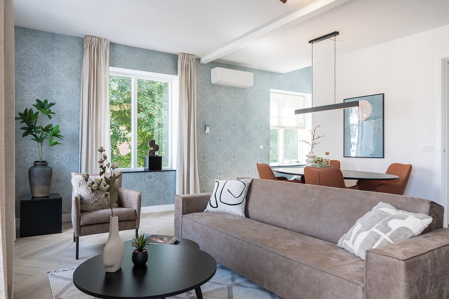 A modern living room with a sofa, armchair, and coffee table, a dining area with chairs and a table, and large windows with curtains.