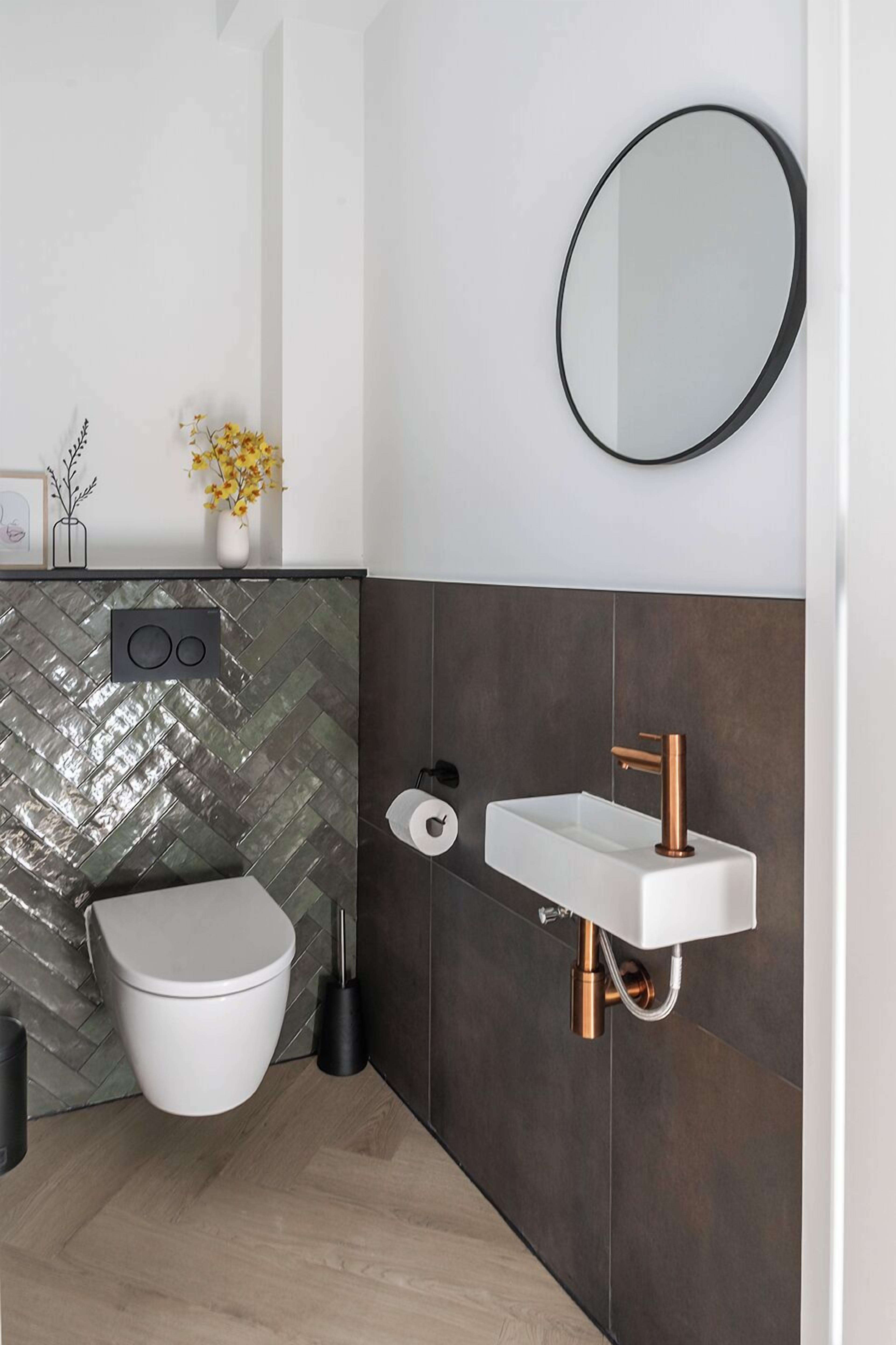A modern bathroom with a white toilet, a white sink with copper fixtures, a herringbone-patterned backsplash, and a round mirror.