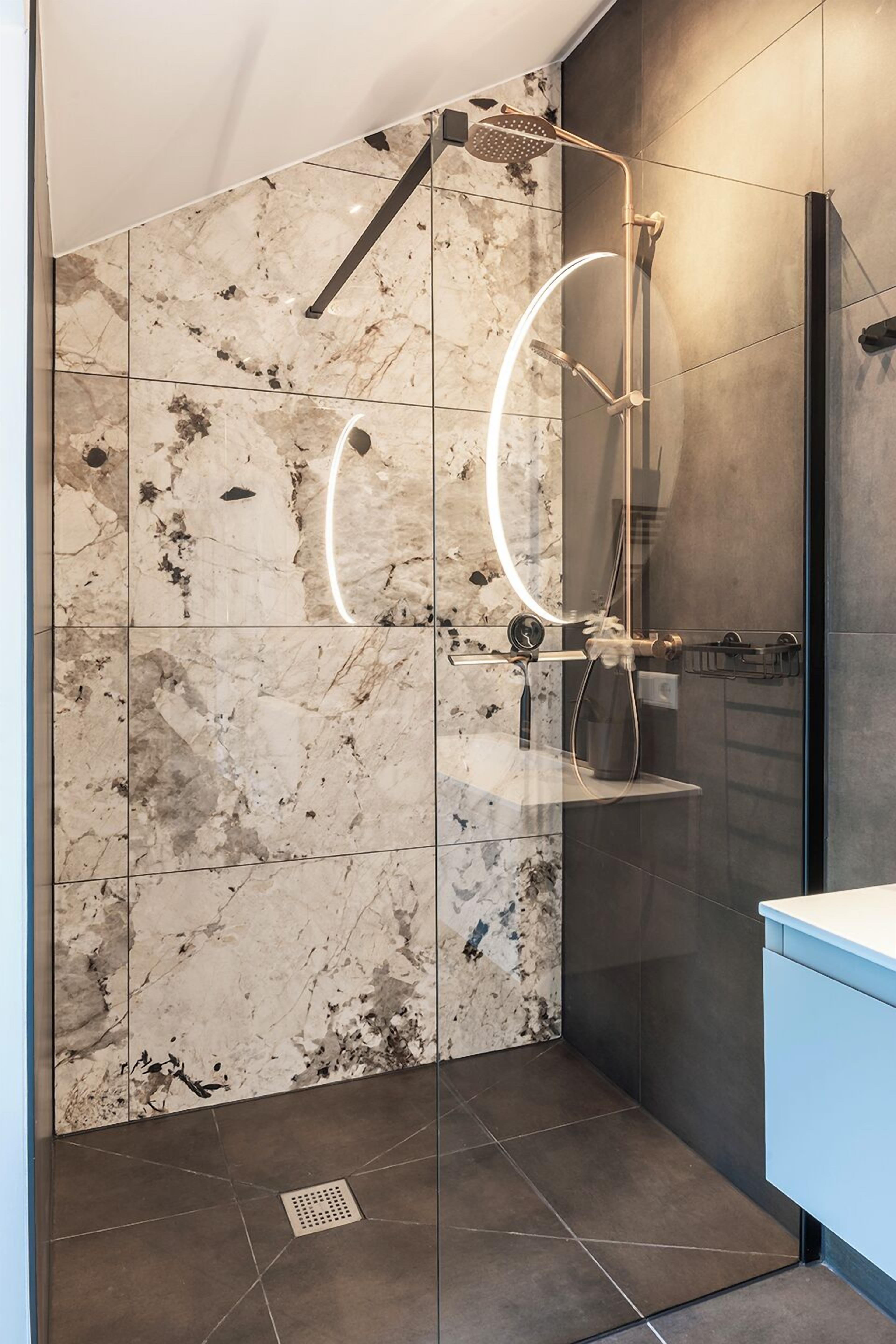 Un baño moderno con ducha envuelta en vidrio, paredes de mármol y suelo oscuro.