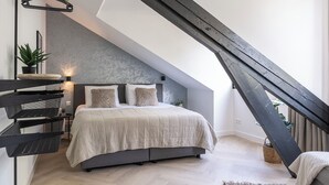 A modern loft bedroom with a sloped ceiling, a bed with a grey headboard, a hanging plant, and a built-in shelving unit.