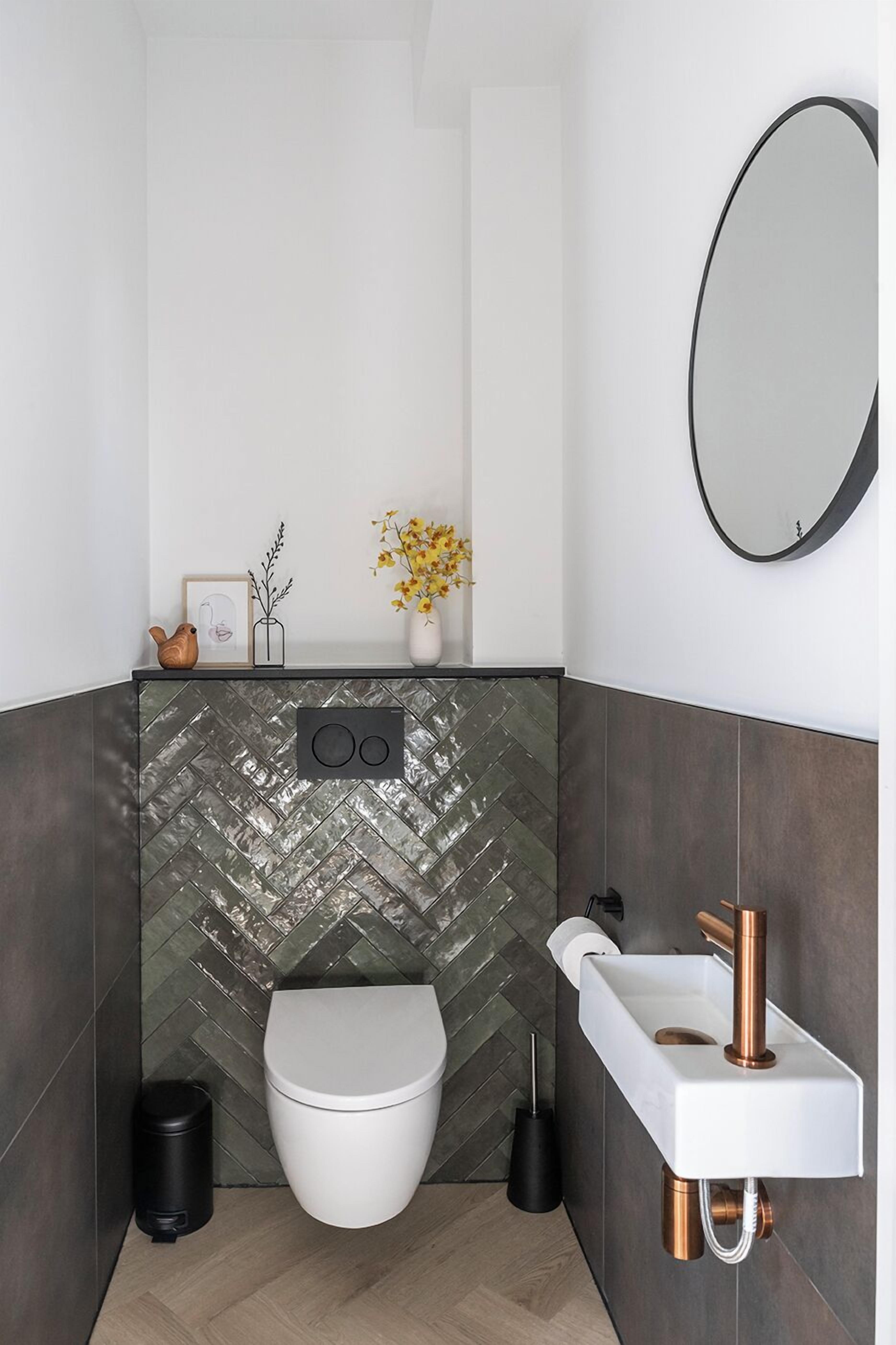 Une salle de bain moderne avec un mur recouvert de carreaux disposés en motif chevron, des toilettes blanches, un lavabo blanc avec des accessoires en cuivre et un miroir rond.