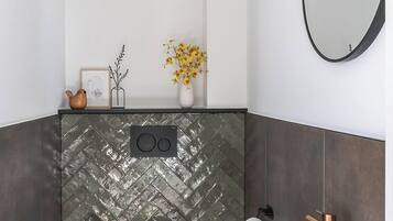 Une salle de bain moderne avec un mur recouvert de carreaux disposés en motif chevron, des toilettes blanches, un lavabo blanc avec des accessoires en cuivre et un miroir rond.
