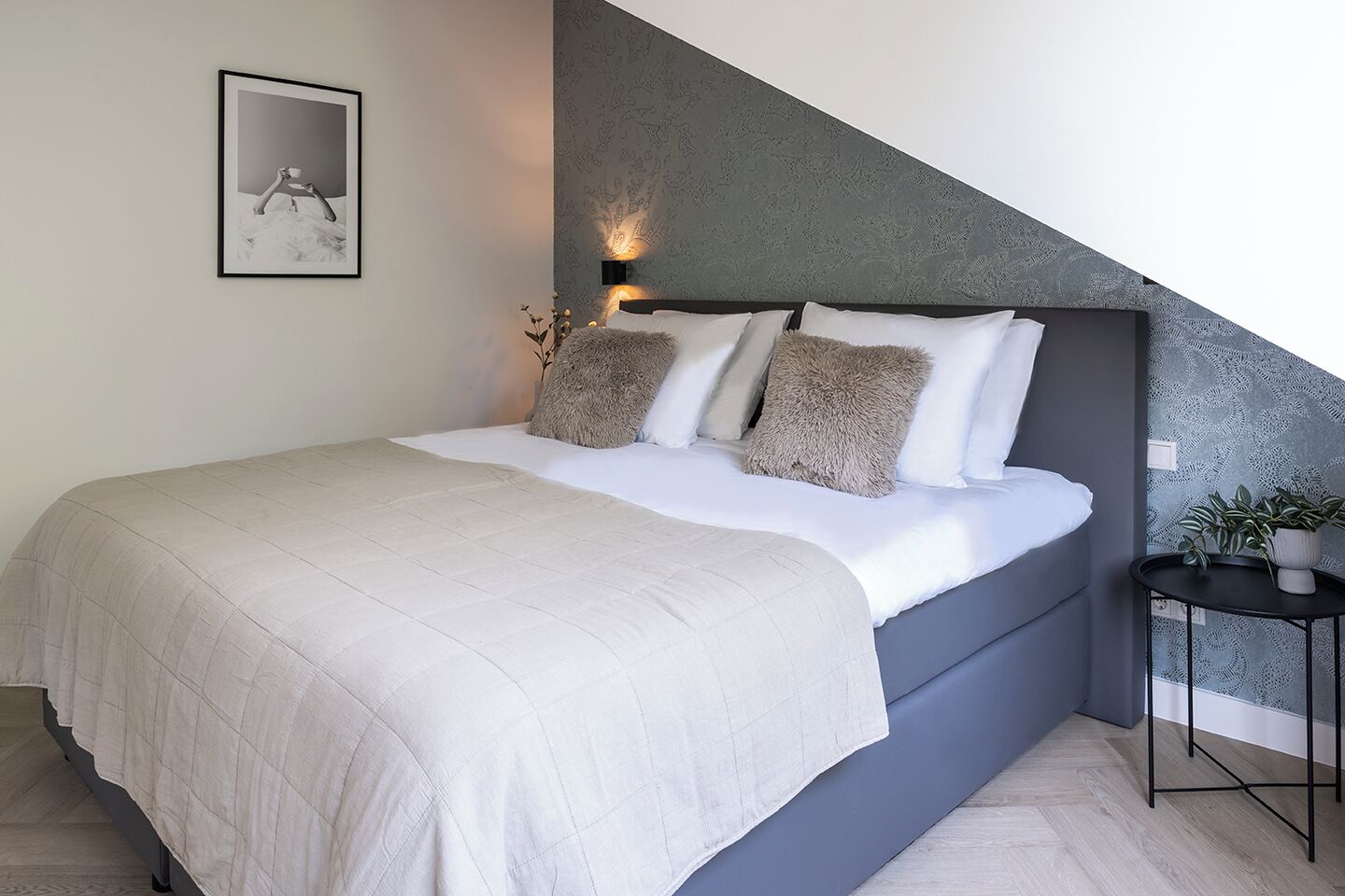 A modern bedroom with a large bed, a headboard with a textured pattern, a bedside table with a plant, and a framed picture on the wall.
