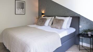 A modern bedroom with a large bed, a headboard with a textured pattern, a bedside table with a plant, and a framed picture on the wall.