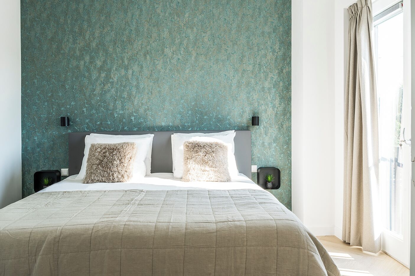 A neatly made bed with a quilted cover, two fluffy pillows, and a headboard against a textured teal wall.