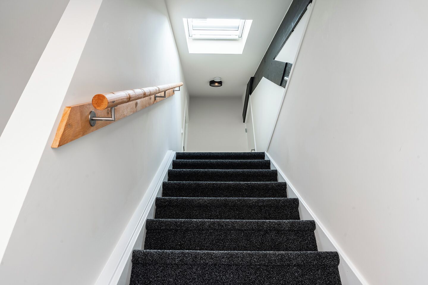 Un escalier avec des marches recouvertes de moquette noire, une rampe en bois et une lucarne au-dessus.