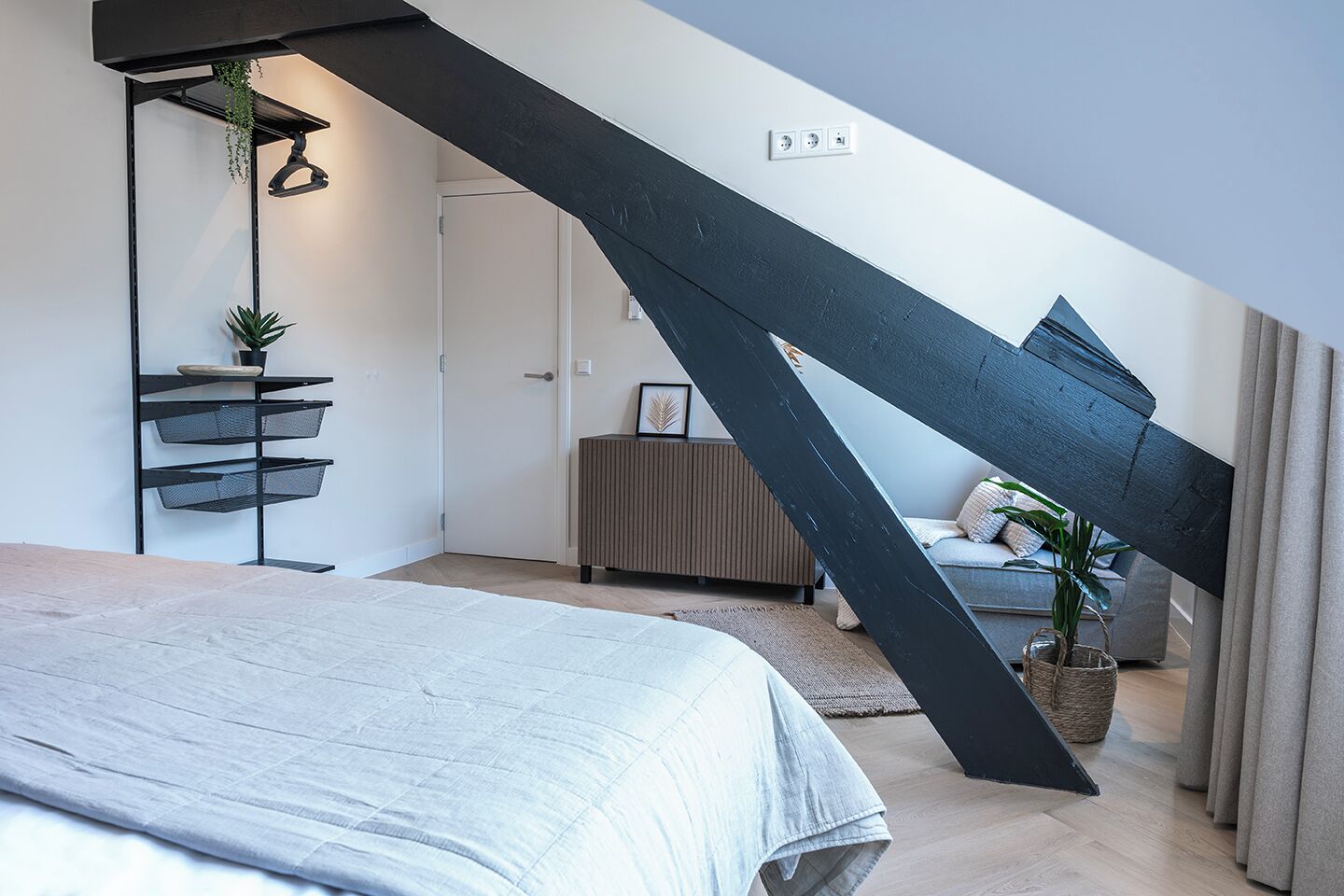 A modern bedroom with a sloped ceiling, a bed, a bedside table with a plant, a radiator, and a door.